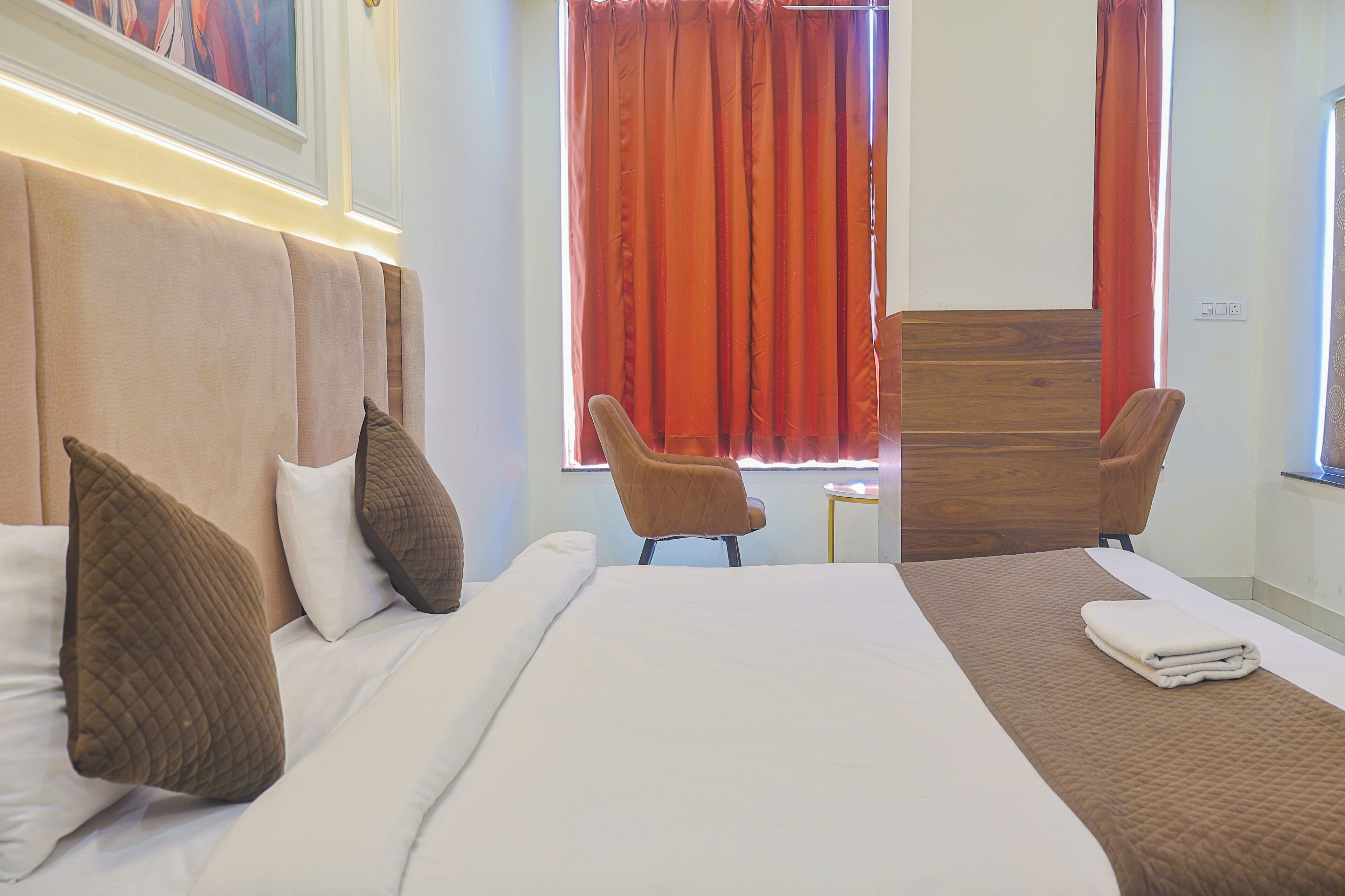 A hotel room with a bed, two chairs, a wooden headboard, and a window with red curtains.