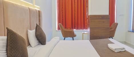 A hotel room with a bed, two chairs, a wooden headboard, and a window with red curtains.