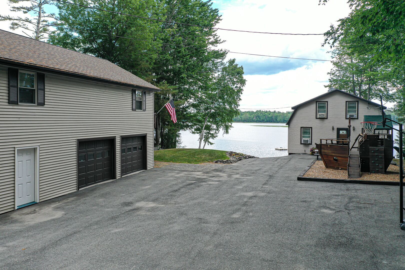 Une maison à deux étages avec un garage, un panier de basketball et une jetée qui s’avance dans un lac.