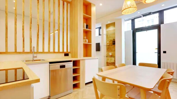 Une cuisine moderne dotée de meubles en bois, d’un lave-vaisselle et d’un coin repas avec une table et des chaises blanches.