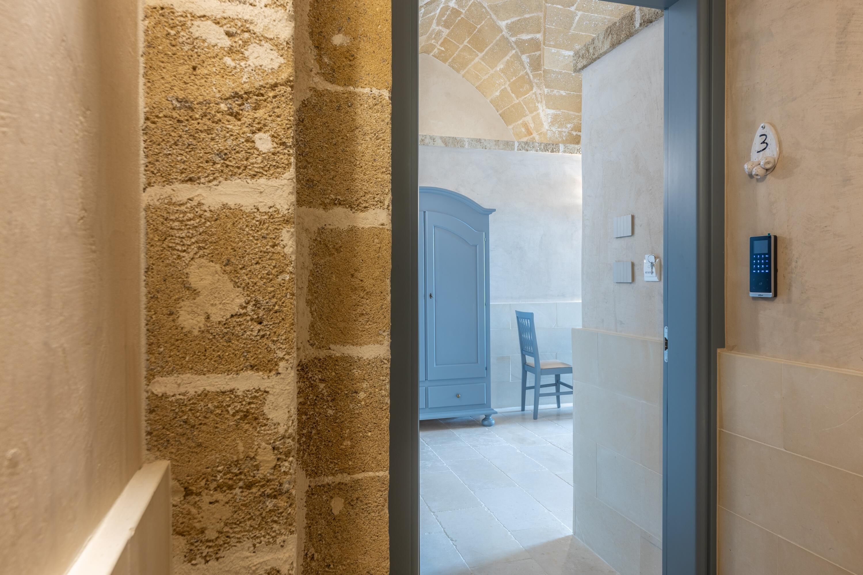 Un couloir aux murs de pierre, une porte bleue et une enseigne portant le numéro 3.
