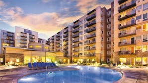 Ein Hotel mit Schwimmbecken, umgeben von Liegestühlen, mehreren Etagen mit Balkonen und einem Sonnenuntergangshimmel.