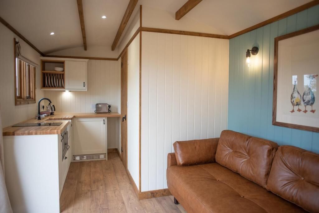 A compact kitchen with wooden cabinets, a built-in dishwasher, and a brown leather sofa.