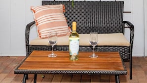 Een rieten bank met een kussen en een gestreept kussensloop, een fles witte wijn en twee wijnglazen op een houten tafel.