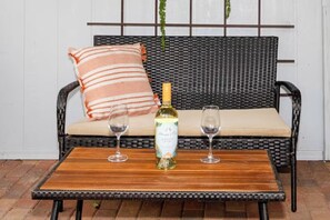 Ein rattangeflochtenes Sofa mit Kissen und einem gestreiften Kopfkissen, eine Flasche Weißwein und zwei Weingläser auf einem Holztisch.