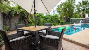 Een terras met een tafel en stoelen onder een zonnescherm, een zwembad met opblaasbaar speelgoed en weelderige begroeiing.