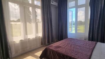 A bedroom with a bed, a ceiling fan, large windows with curtains, and a view of the outdoors.