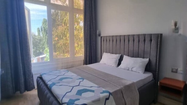 A bedroom with a large bed, a window with curtains, a bedside table, and a view of trees outside.