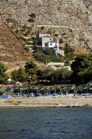 A coastal property with a stone house, beach umbrellas, and a rocky hillside. - VILLA ATHINA (Hydra)