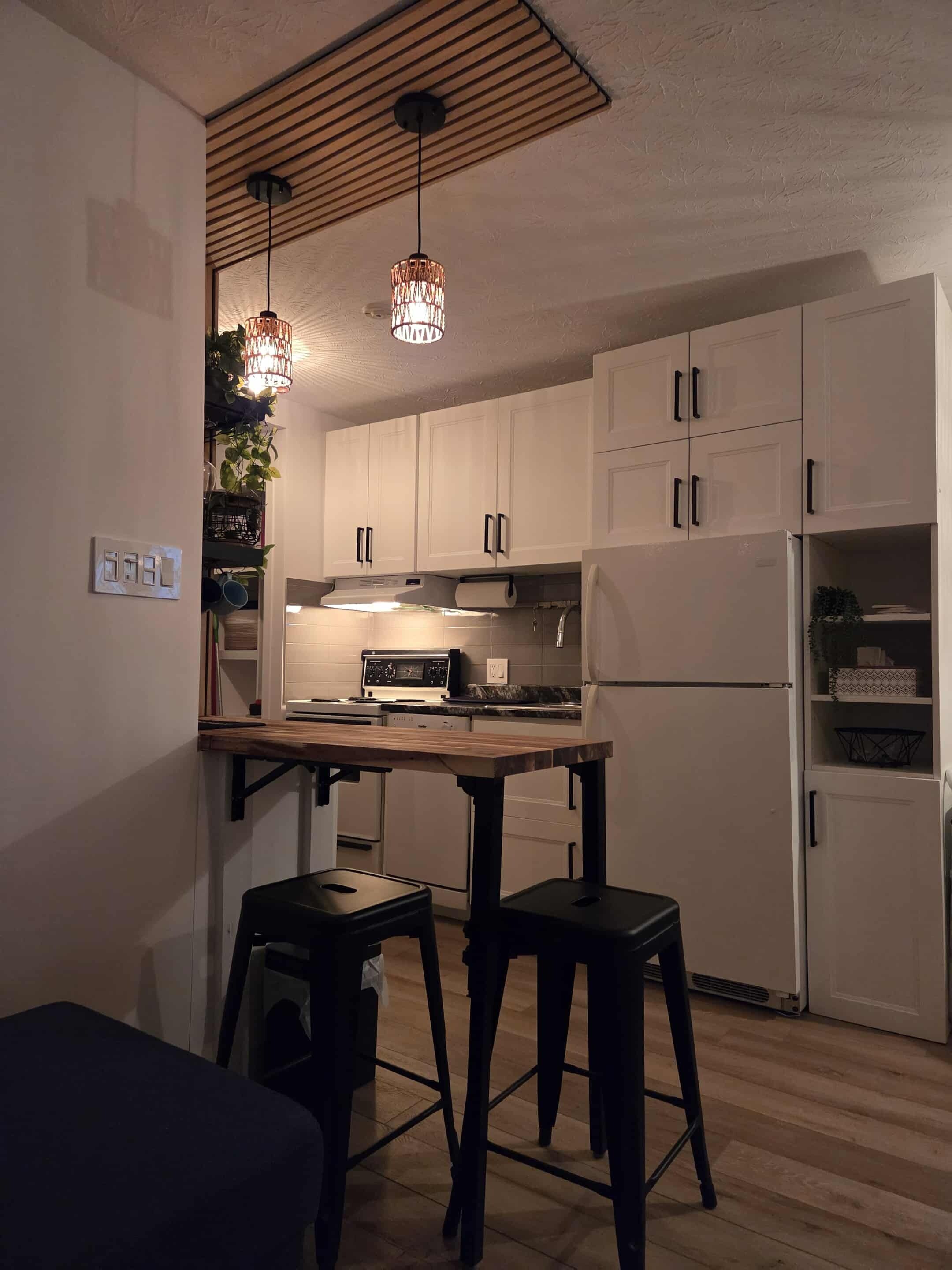 A compact kitchen with white cabinets, a wooden countertop, two stools, and pendant lighting.