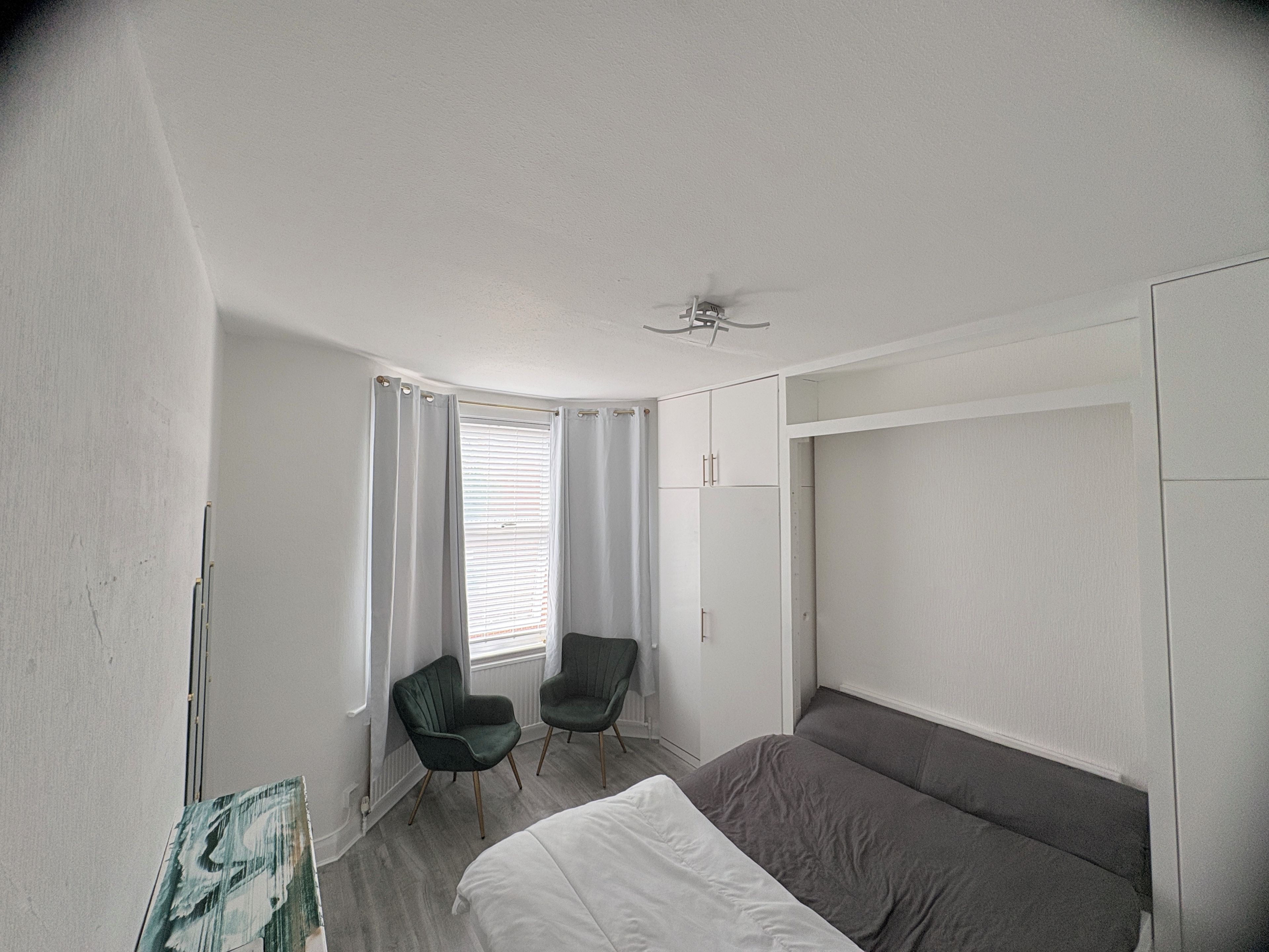 A bedroom with a bed, two chairs, a window with curtains, a ceiling fan, and a wardrobe.