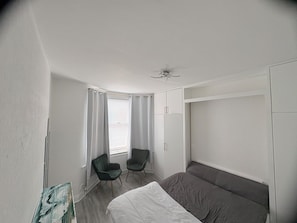 A bedroom with a bed, two chairs, a window with curtains, a ceiling fan, and a wardrobe. - Chill Spot (London)
