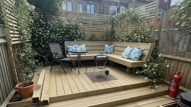 Eine Holzterrasse mit einer Bank und einem Tisch, umgeben von einem Holzzaun und Kübelpflanzen.
