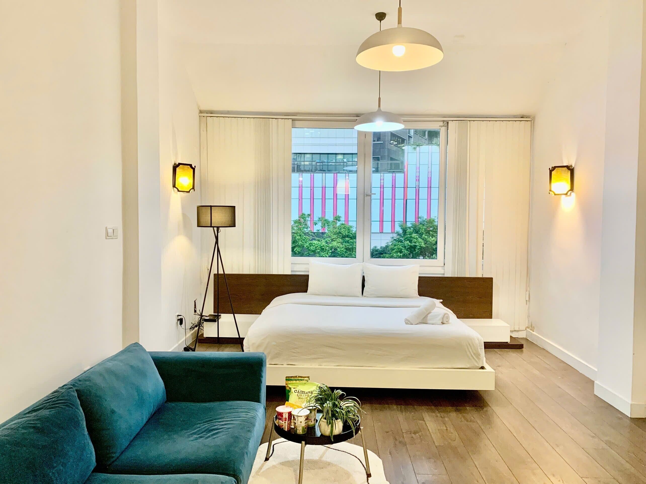 Ein modernes Hotelzimmer mit einem großen Bett, einem blauen Sofa, einem kleinen Tisch mit Snacks und Blick auf ein Gebäude mit pinken und grünen Elementen vor dem Fenster.