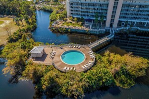 Eine Vogelperspektive zeigt ein Resort mit einem runden Pool, umgeben von einer Holzterrasse, einer Brücke und üppiger Vegetation.