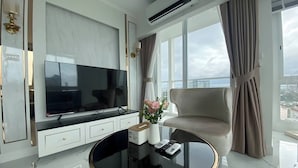 A modern hotel room with a large flat-screen TV, a round glass coffee table, a comfortable armchair, and a view of the city through large windows. - 2 Bed Room Sea View Great Place 2 (Pattaya)