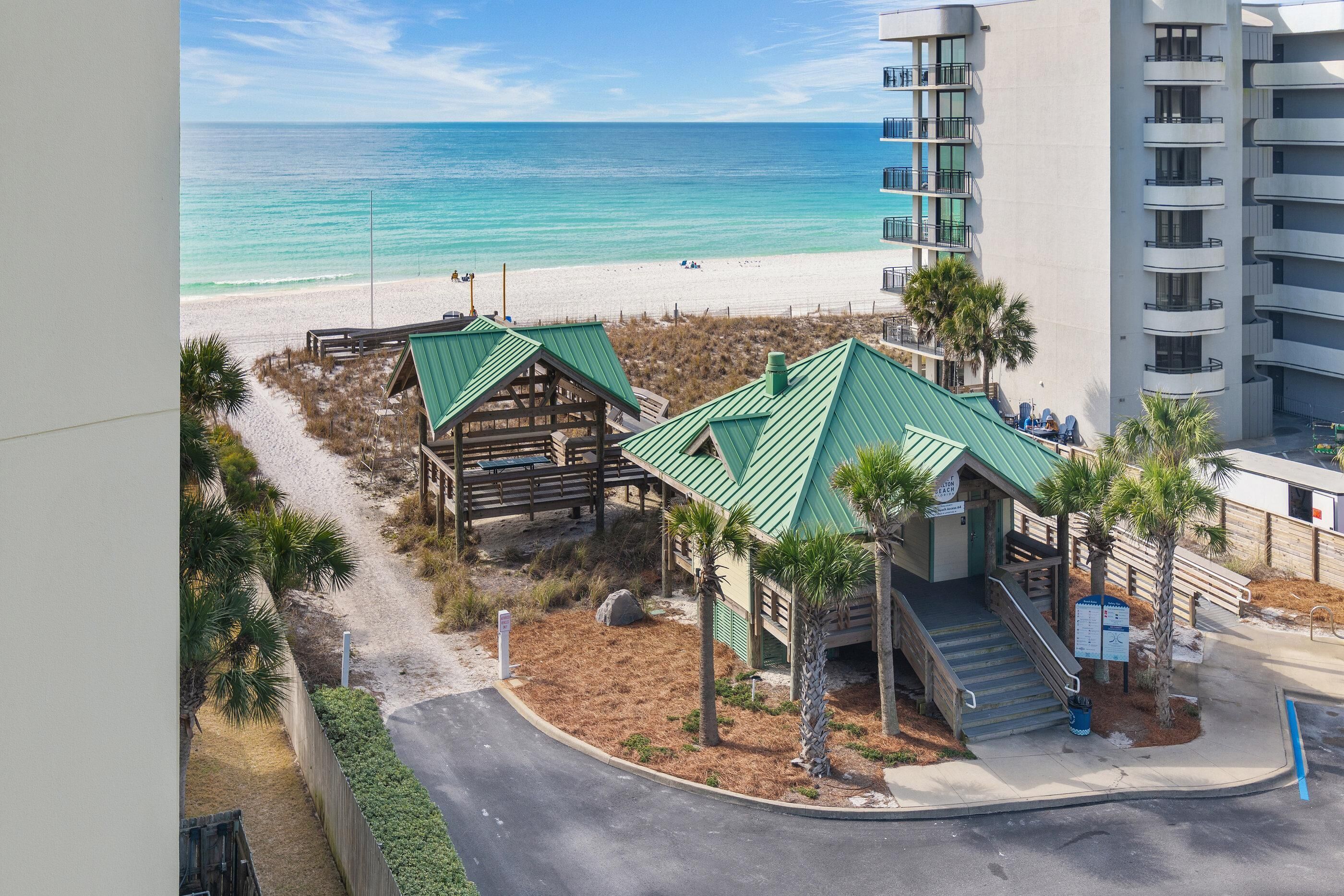 Un hotel frente a la playa con estructura de madera y techo verde, junto a una playa de arena y el océano.