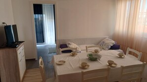 Ein Zimmer mit Esstisch für vier Personen, einer Couch, einem Fernseher auf einem Schrank und einem Fenster mit Vorhängen.