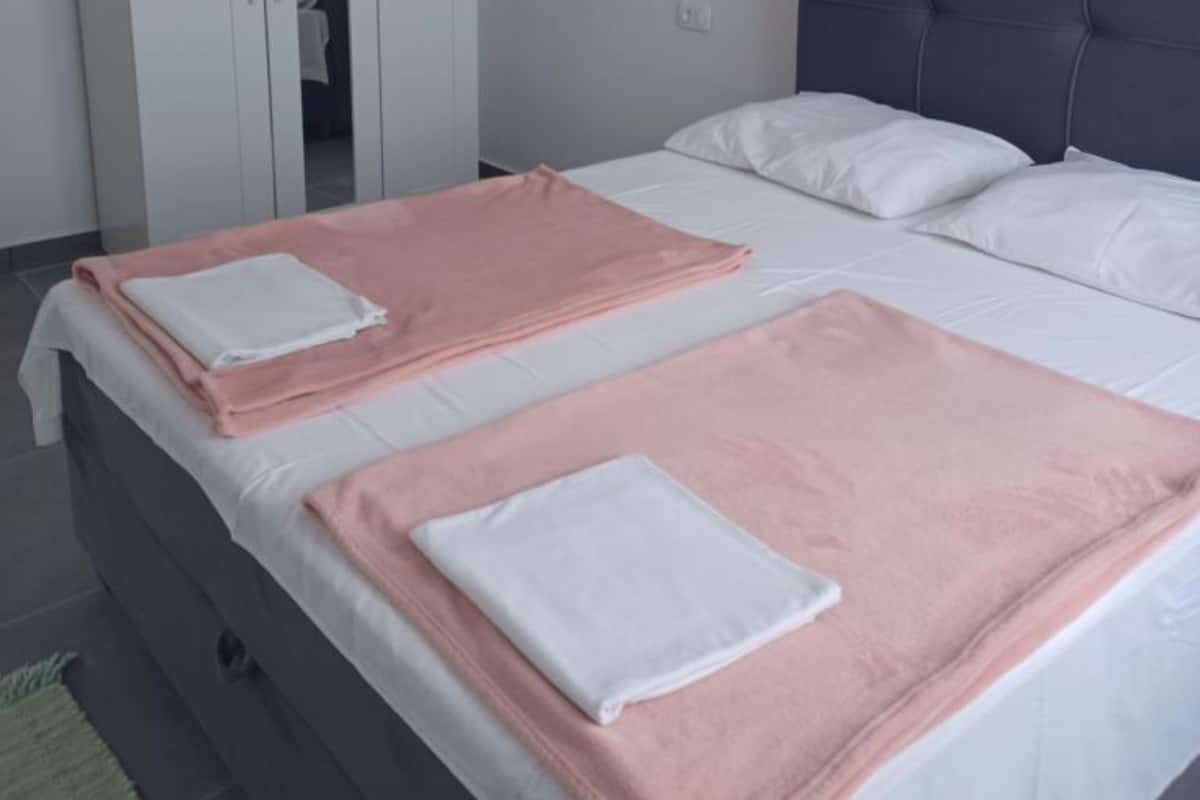 Een netjes opgemaakt bed met witte en roze linnens, een grijs hoofdbord en een zichtbare garderobe op de achtergrond.