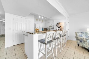 A modern kitchen with white cabinets, a granite countertop, and a dining area with white chairs. - Emerald Beach 1928 (Panama City Beach)