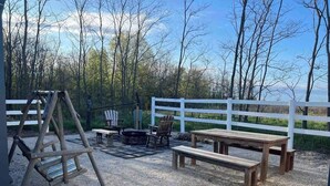 Un espace extérieur aménagé en patio, avec une balançoire en bois, un foyer, des bancs en bois et une clôture blanche.
