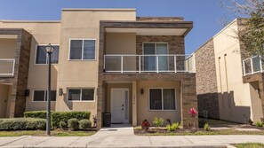 A modern two-story apartment building with a balcony, a front door, and a small garden area with plants. - Incredible 4BD Luxury Home with BBQ and Lake View (Kissimmee)