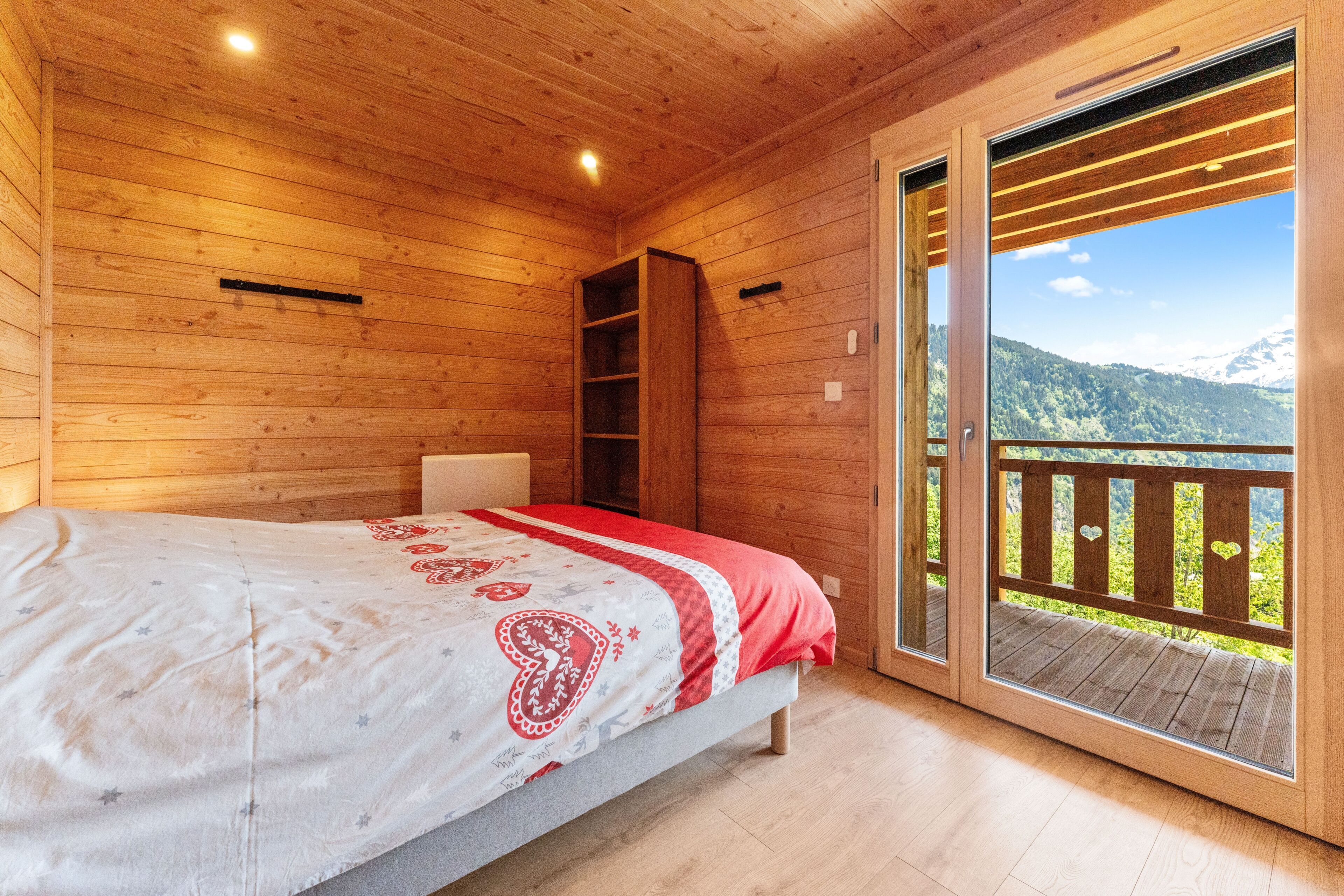Una habitación de madera con una cama, un estante y una puerta corredera que da a un balcón con vista a la montaña.