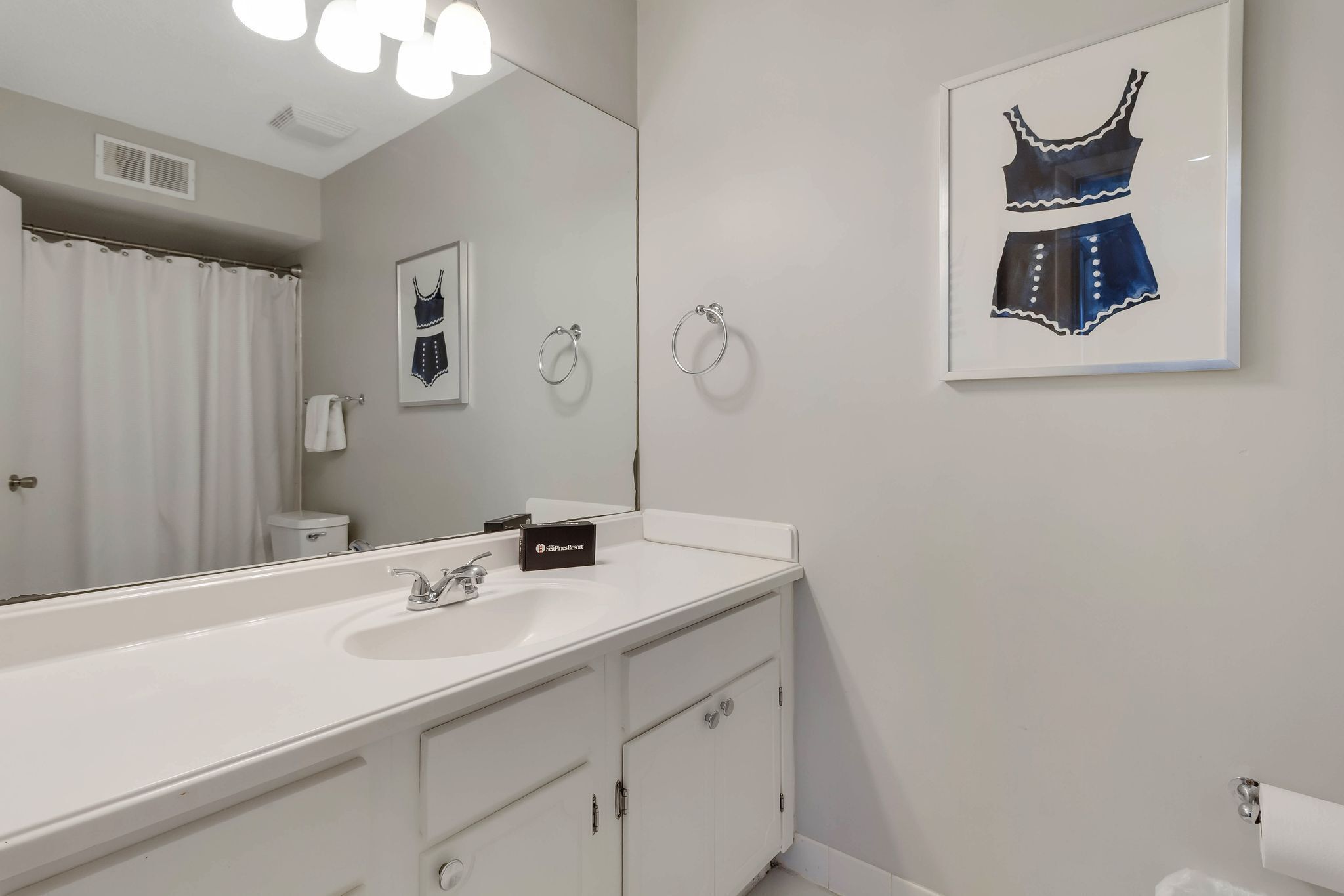A modern bathroom with a large mirror, white vanity, and a framed artwork of a dress on the wall.