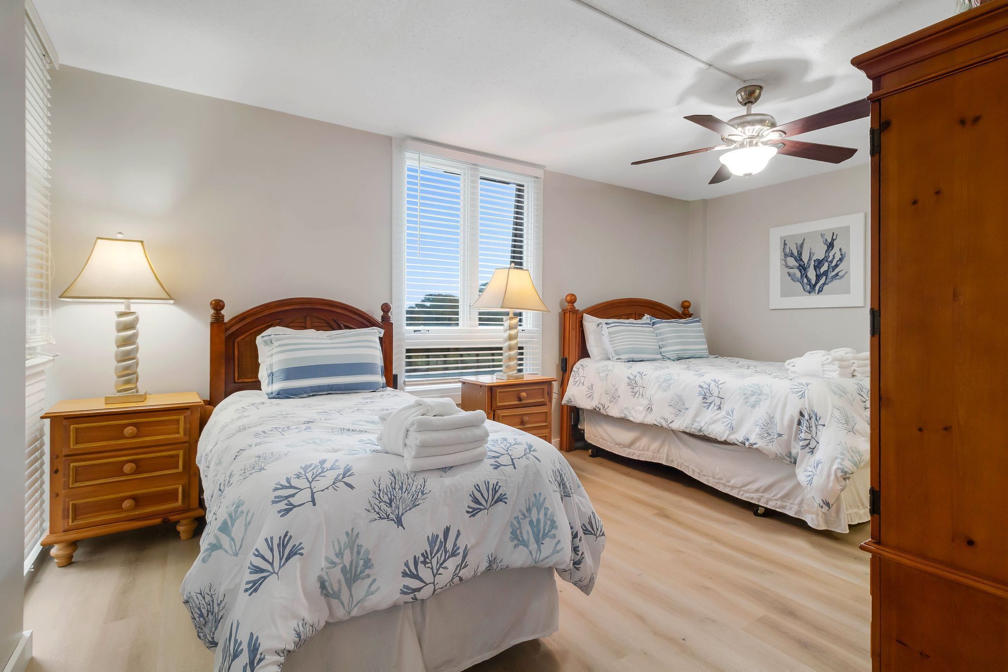 Une chambre avec deux lits, un ventilateur de plafond, une fenêtre avec des stores et une armoire en bois.