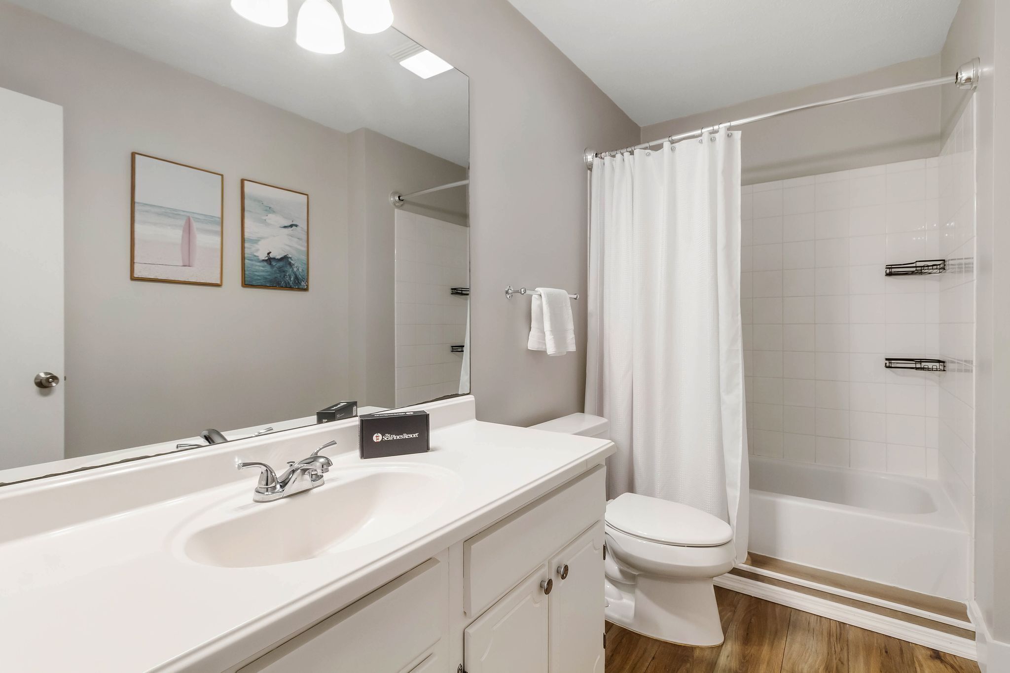 A modern bathroom with a white sink, toilet, and shower. There are framed pictures on the wall and a towel rack.