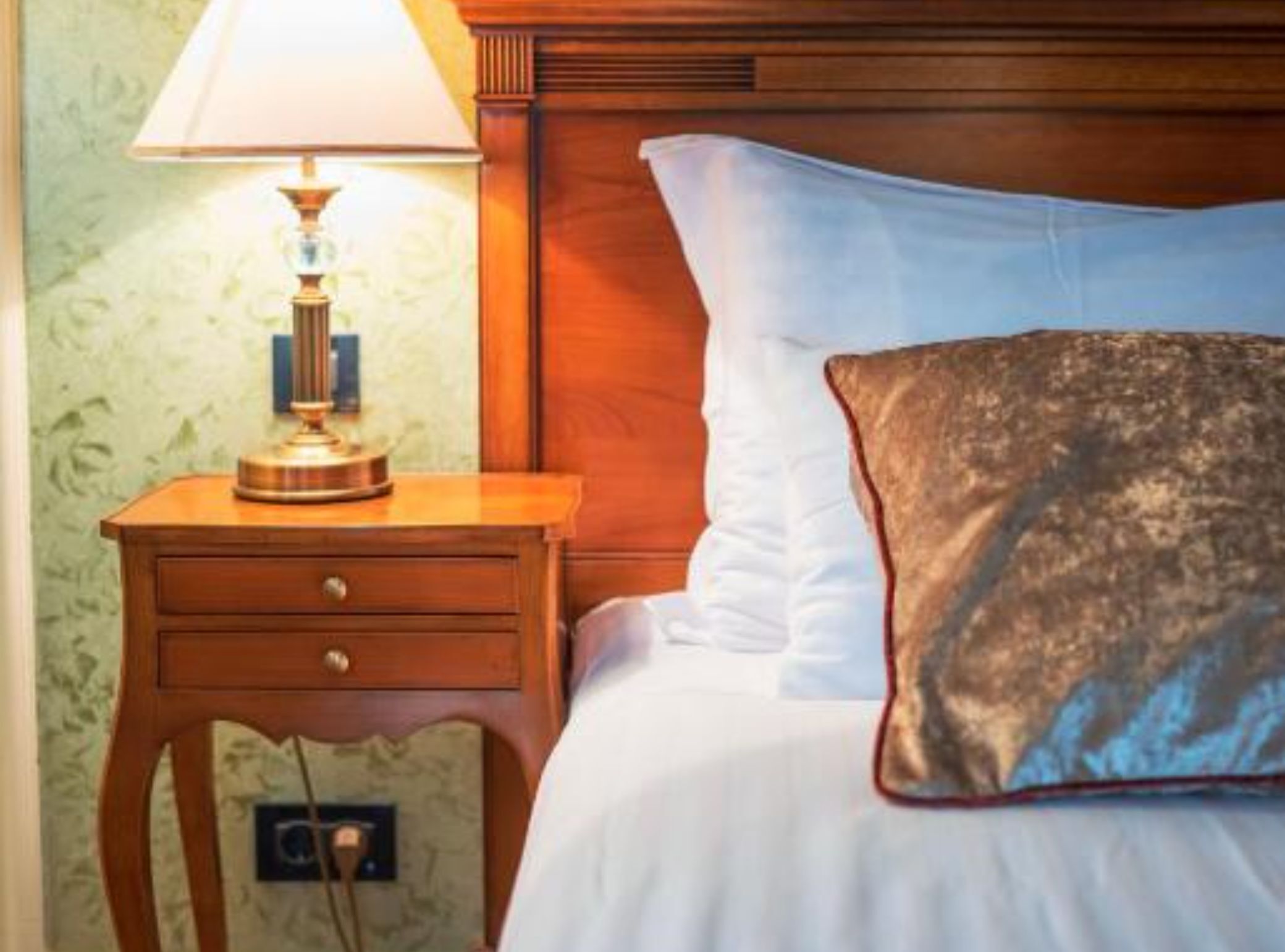 Una habitación de hotel con cabecera de madera, una mesita de noche con lámpara, una cama individual con ropa blanca y un cojín decorativo estampado.