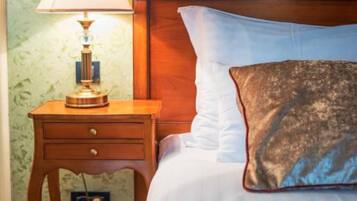 Una habitación de hotel con cabecera de madera, una mesita de noche con lámpara, una cama individual con ropa blanca y un cojín decorativo estampado.