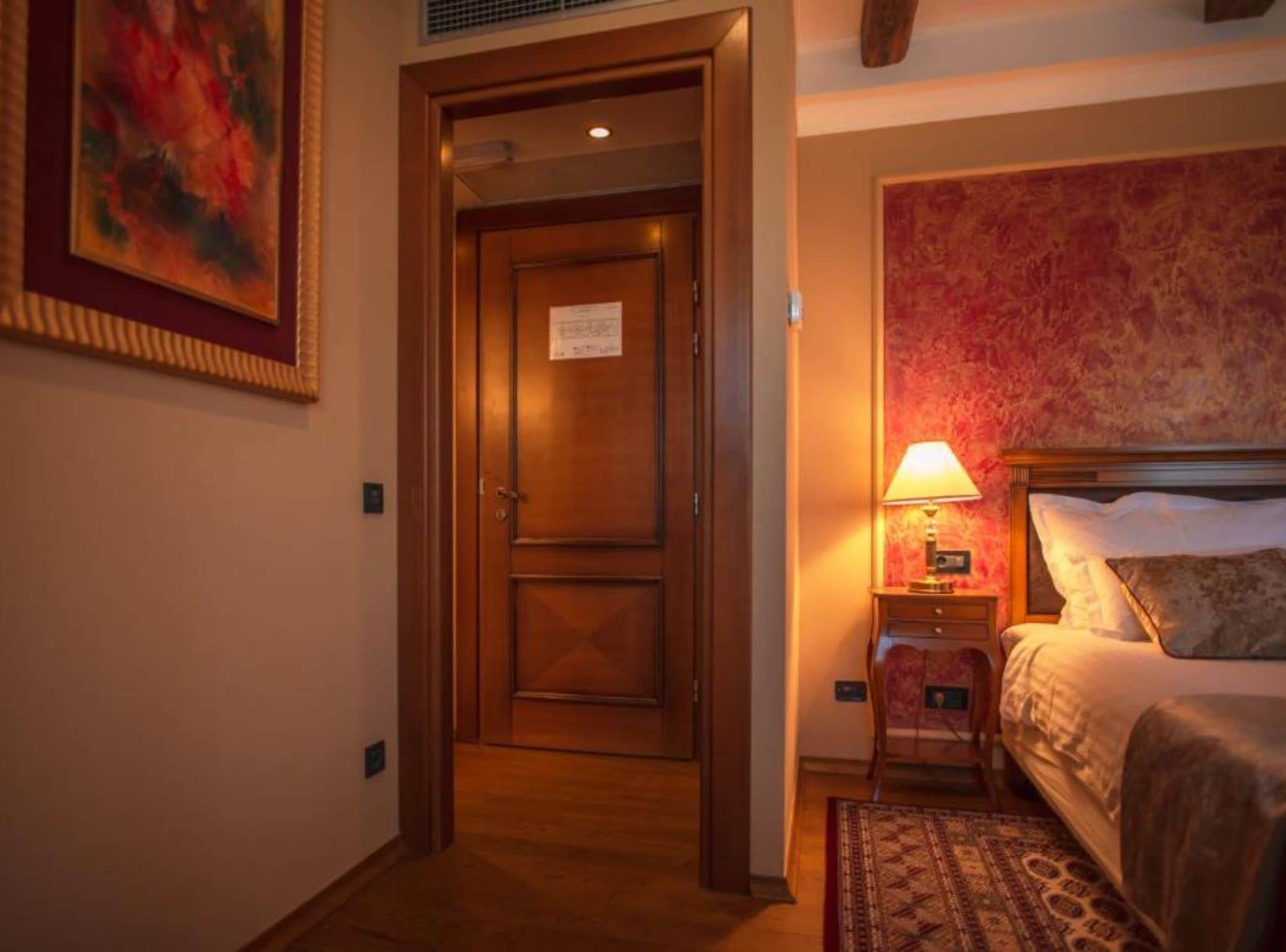 A hotel room with a wooden door, a bed with a patterned headboard, a nightstand with a lamp, and a small rug.