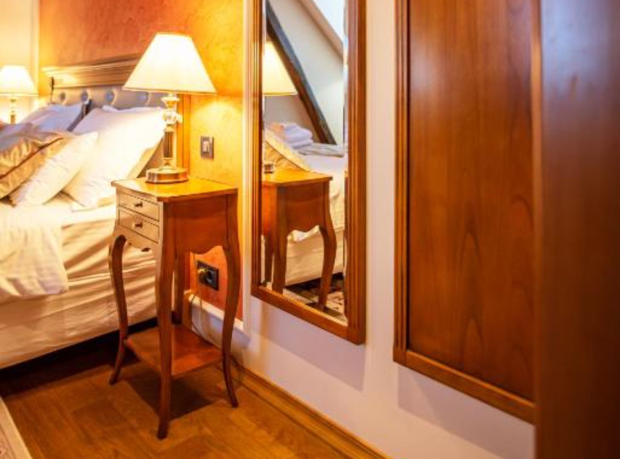 Habitación de hotel con cama, lámparas de noche, mesita de noche de madera, espejo y puerta de madera.