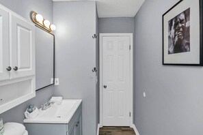 Une salle de bain avec un meuble-lavabo blanc, un miroir avec éclairage intégré, une porte et un tableau encadré au mur.