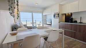 A modern kitchen with a built-in oven, a coffee machine, and a sink. There is a dining area with a table and chairs, and a view of the beach through large windows. - Beachfront Studio w/Balcony & Family Amenities (Knokke-Heist)