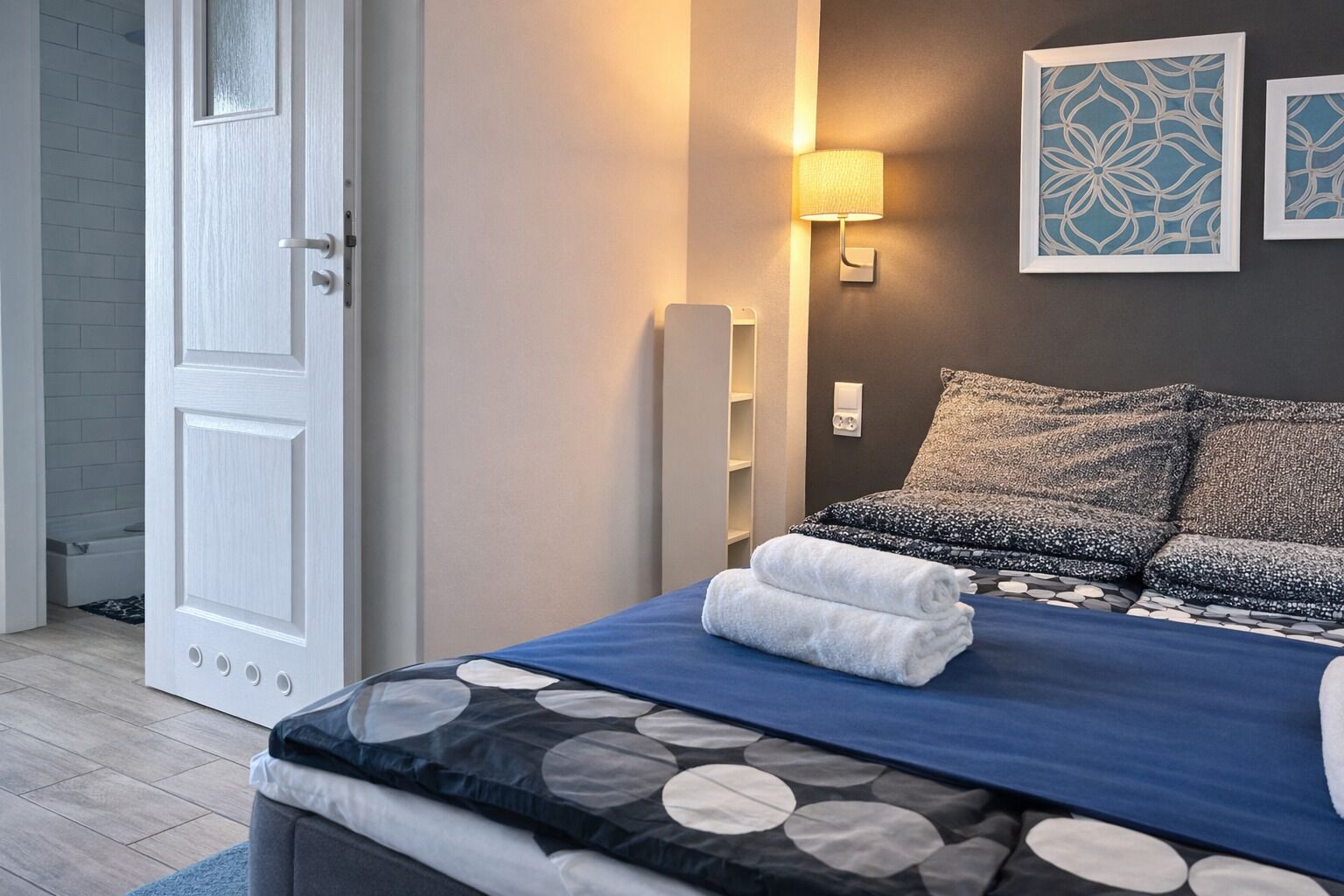 Un lit bien fait, recouvert d’une couvre-lit à motifs bleus et blancs, avec deux oreillers et une serviette pliée posée sur le lit. Une lampe fixée au mur et deux œuvres d’art encadrées sont accrochées au-dessus du lit.