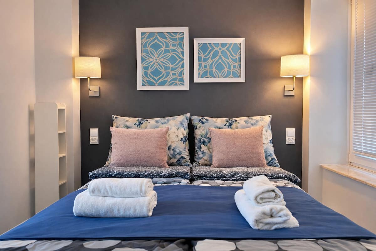 Una cama bien hecha con almohadones estampados con motivos florales, dos lámparas de pared y dos cuadros enmarcados.