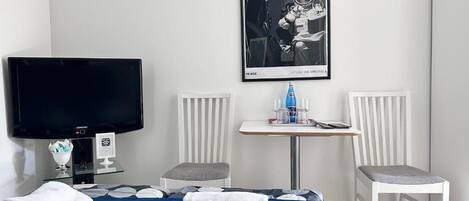 Een hotelkamer met een bed, een televisie, een kleintje tafeltje met een fles, twee stoelen, en een ingelijst kunstwerk aan de muur.