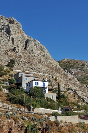 A two-story stone house with blue shutters, situated on a hillside with rocky terrain and sparse vegetation. - VILLA FIVOS (Hydra)