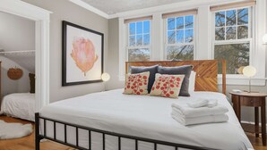 A bedroom with a large bed, a wooden headboard, a framed artwork on the wall, and a window with a view of trees. - Charming 4BR Bungalow - Steps to Clarendon METRO (Arlington)