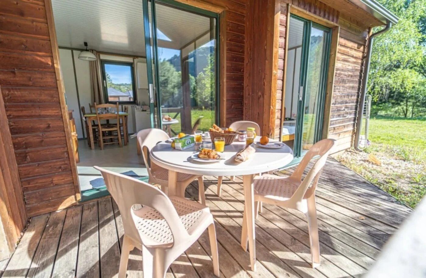 Une terrasse en bois avec une table ronde et des chaises, le petit-déjeuner est servi, et la vue donne sur un paysage verdoyant.