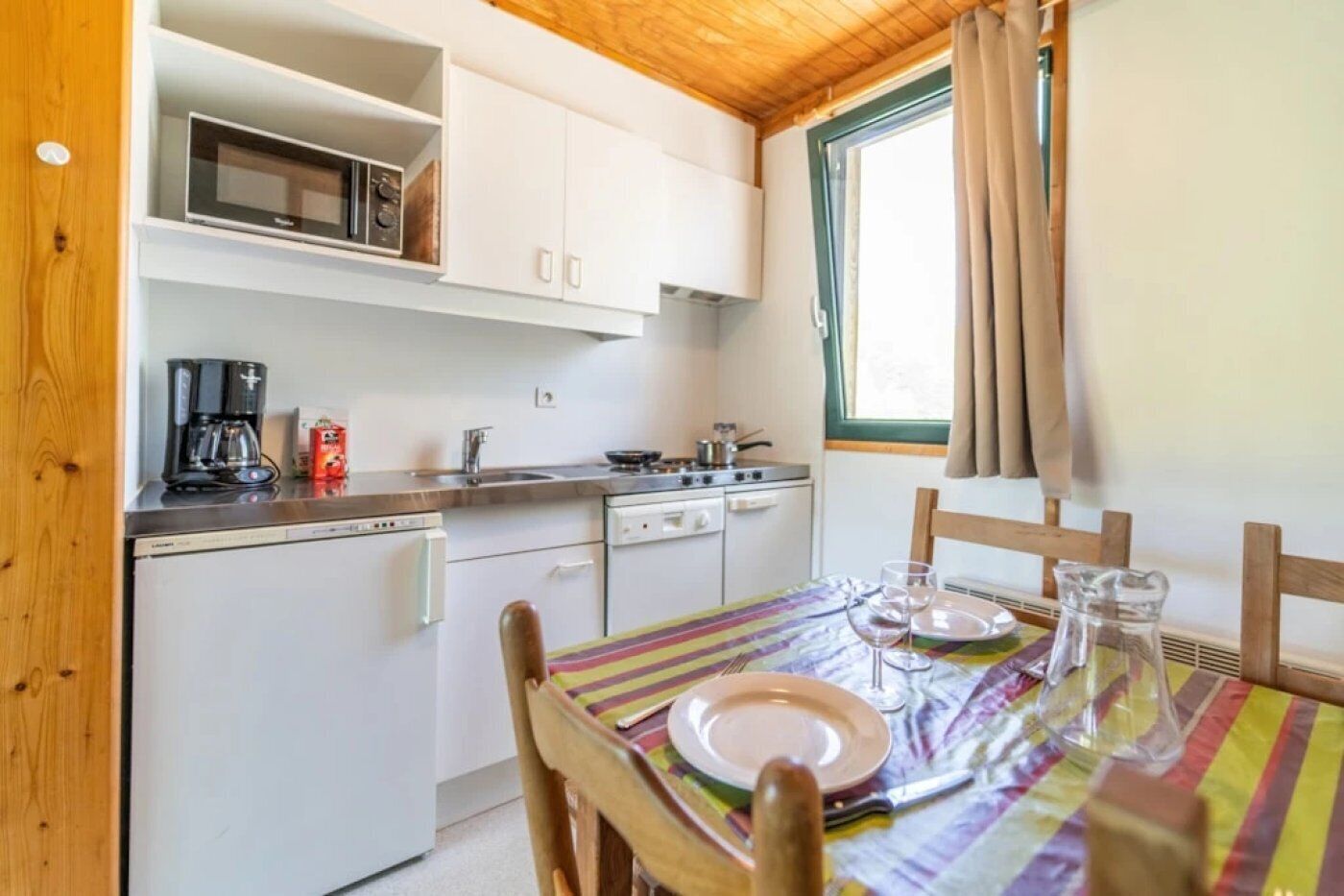 Une cuisine compacte avec des meubles blancs, un four à micro-ondes, une machine à café et une table à manger, ainsi que de l’assiette et des verres.