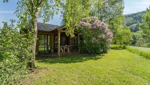 A wooden cabin with a covered porch, surrounded by lush greenery and flowering bushes.