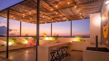 Una terraza al aire libre con techo de paja, luces colgantes, asientos y vistas a la ciudad al atardecer.