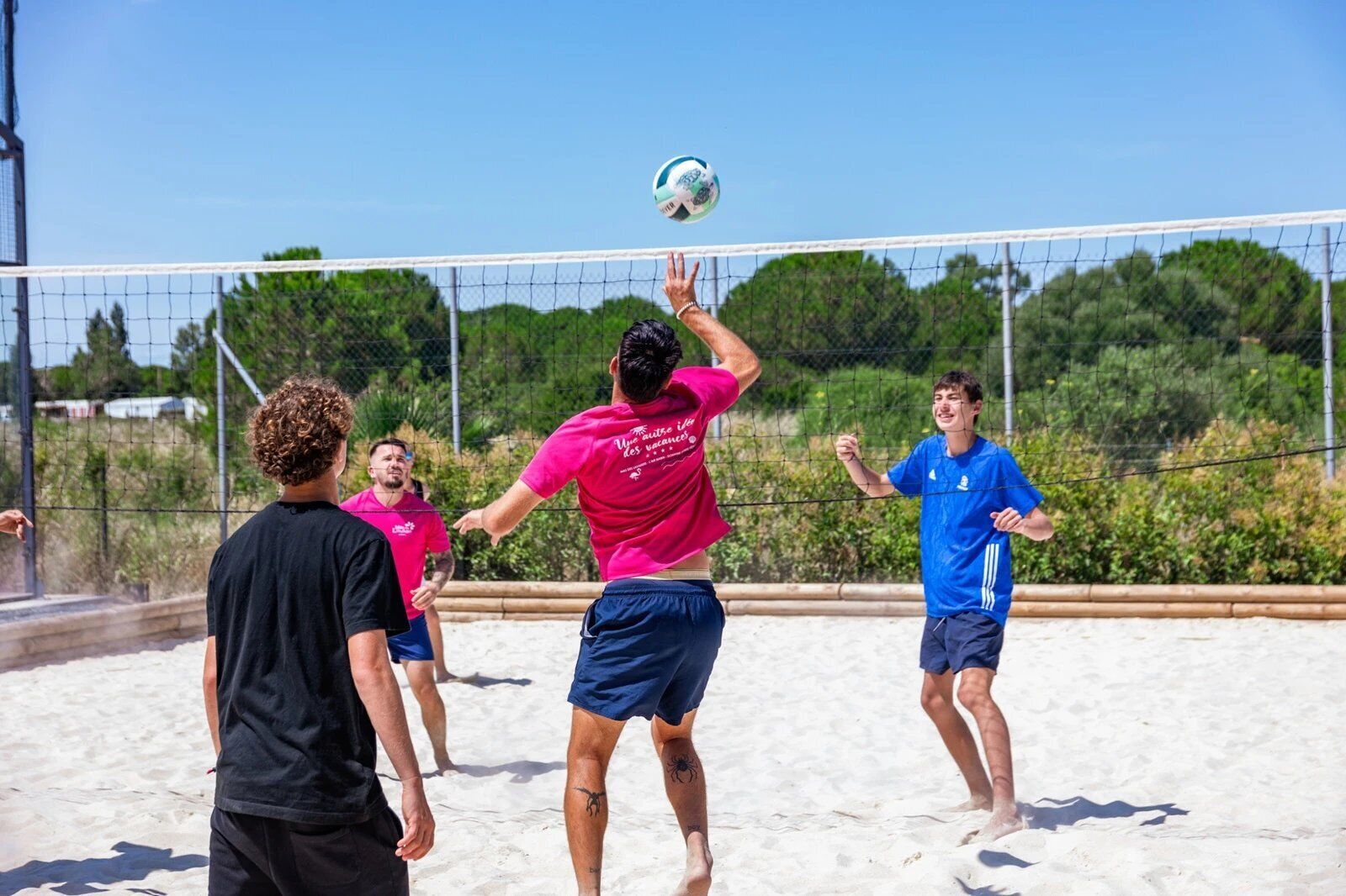 Un groupe de personnes joue au volleyball de plage sur un terrain sablonneux, délimité par un filet, et entouré de verdure sous un ciel dégagé.