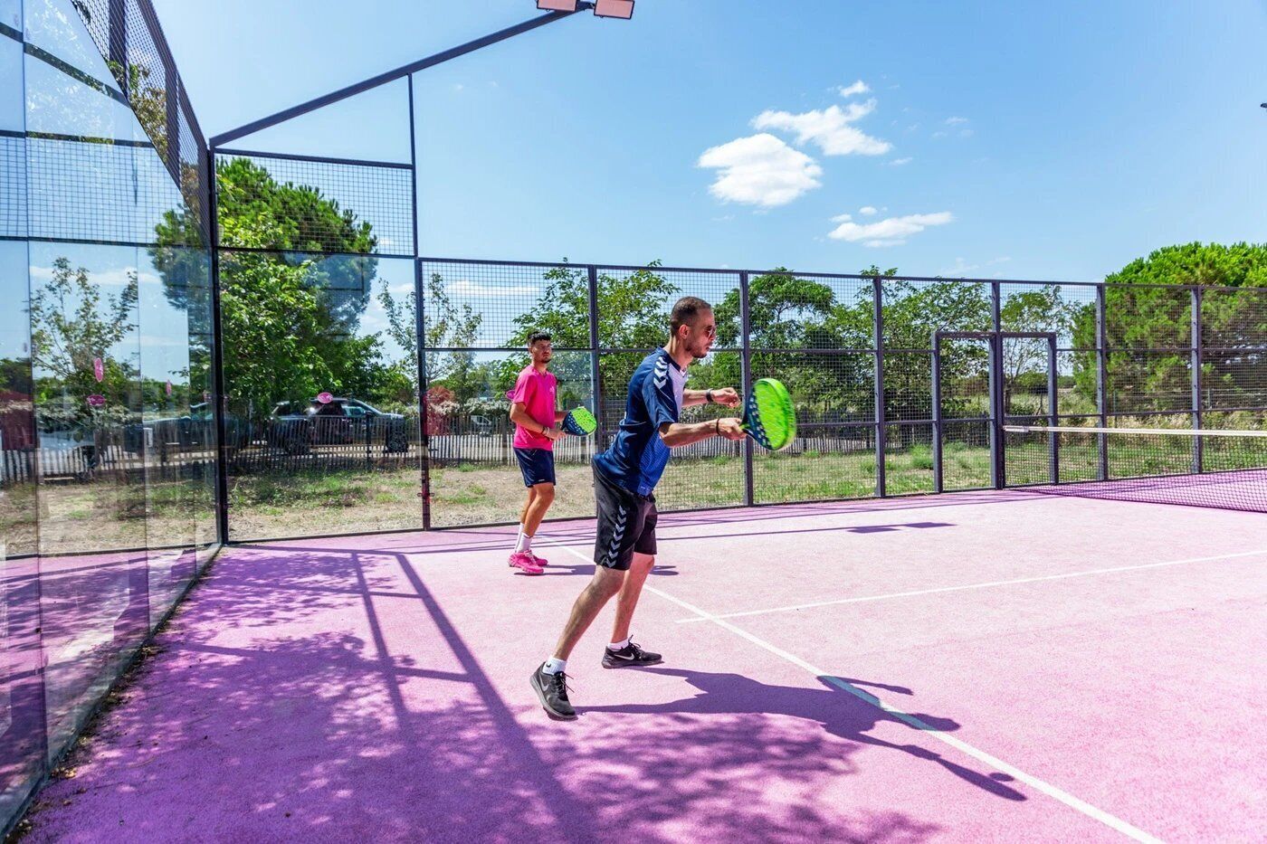 A tennis court with two players in action, surrounded by a fence and trees.