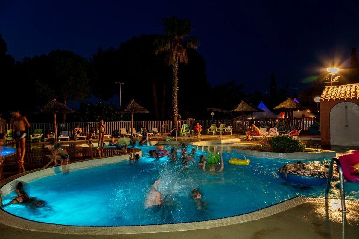 A swimming pool area with people swimming and lounging, surrounded by palm trees and umbrellas.