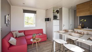 A compact living space with a red sofa, a small round table with a plant, a kitchen area with white cabinets, and a dining table set for four. - ZEcamping 4* - espace aquatique de 2000m2 - proche cap d'Agde (Bessan)