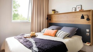 A modern bedroom with a large bed, a wooden headboard, two bedside lamps, a window with curtains, and a shelf with decorative items. - ZEcamping 4* - espace aquatique de 2000m2 - proche cap d'Agde (Bessan)